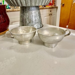 May include: Two silver metal cups with a flared lip and a handle. The cups are sitting on a white surface.