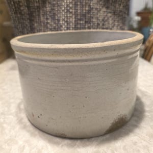 May include: A cylindrical, off-white stoneware pot with a slightly flared rim. The exterior features horizontal striations and subtle colour variations, suggesting a vintage or rustic aesthetic. The rim has a light brown edge.