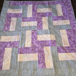 May include: A handmade quilt with a geometric pattern. The quilt features a combination of light blue, white, and purple fabrics. The purple sections have a floral pattern. The quilt is likely made of cotton and is suitable for a bed or as a decorative throw.