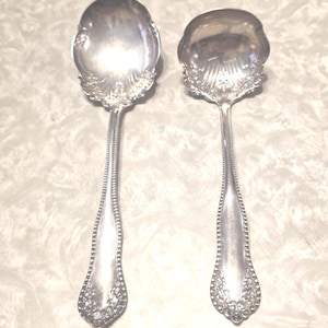 May include: Two antique silver serving spoons. The spoons have ornate floral detailing around the bowl and along the handle. The handles are long and slender, with a beaded design. The spoons are likely for serving food.