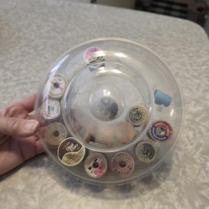 May include: A clear plastic sewing kit containing several spools of thread in various colors. The spools are arranged inside the circular container. A small blue thimble is also visible.
