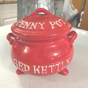 May include: A bright red ceramic pot with a lid. The lid has a handle and the words "PENNY POT" in white lettering. The pot's body has the words "RED KETTLE" in white lettering. The pot has three small feet.