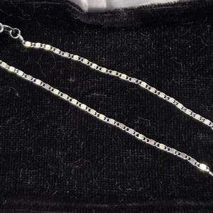 May include: A delicate silver chain necklace with a repeating oval link design. The necklace is displayed on a black velvet surface, highlighting its shine and intricate pattern. The clasp is visible at the end of the chain.