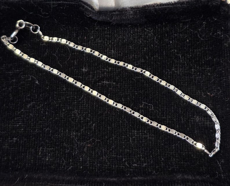 May include: A delicate silver chain necklace with a repeating oval link design. The necklace is displayed on a black velvet surface, highlighting its shine and intricate pattern. The clasp is visible at the end of the chain.