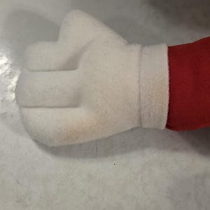 Peut inclure: Une main blanche en peluche en forme de poing, avec un poignet blanc et du tissu rouge visible. La main est faite d'un mat&eacute;riau doux et textur&eacute;.