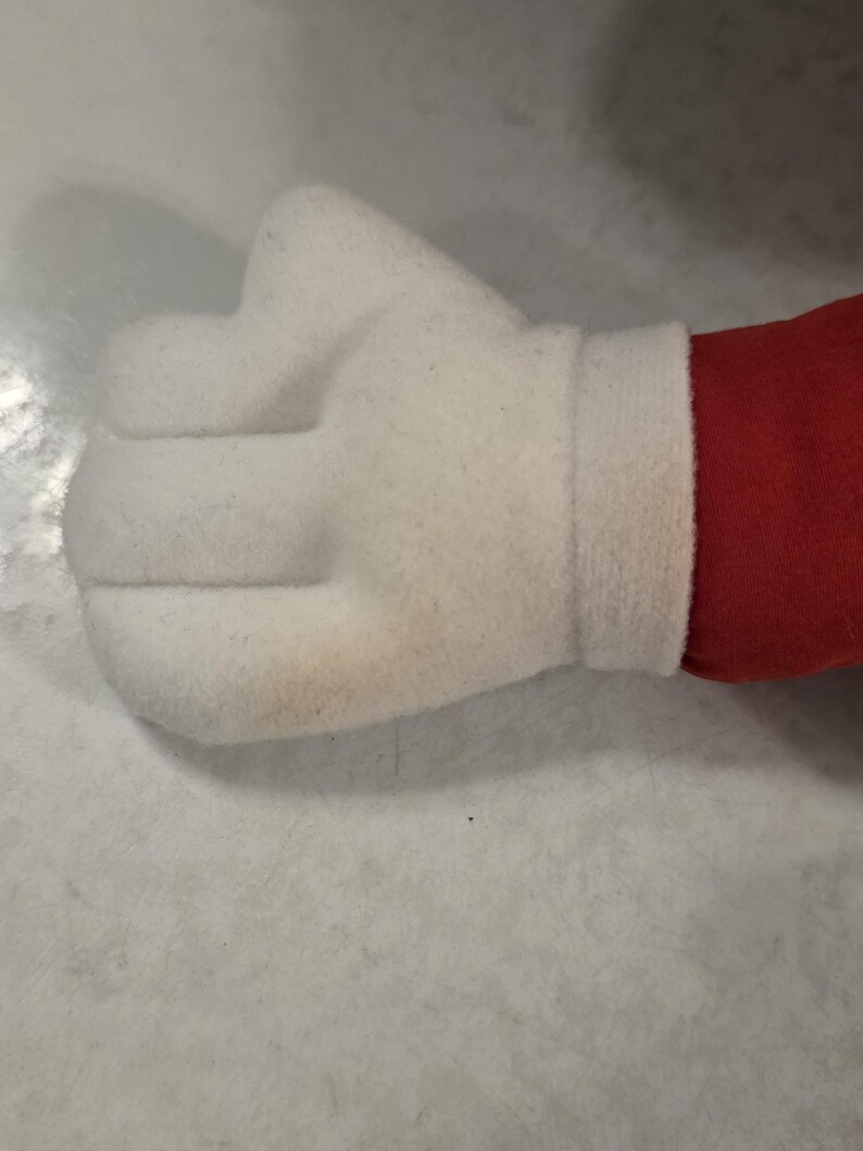 Peut inclure: Une main blanche en peluche en forme de poing, avec un poignet blanc et du tissu rouge visible. La main est faite d'un mat&eacute;riau doux et textur&eacute;.