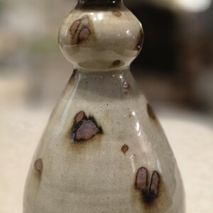 May include: A ceramic bud vase with a bulbous neck and a tapered body. The piece features a glossy, off-white glaze with scattered, dark brown, irregular spots and a smooth, rounded rim. The base shows a hint of unglazed, reddish-brown clay.