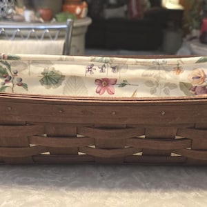 May include: A rectangular brown woven basket with a floral patterned fabric lining. The basket is made of woven wood strips and has a rectangular shape. The fabric lining features a pattern of flowers and leaves.