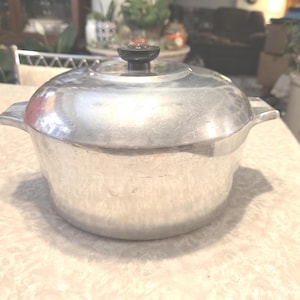 May include: A vintage aluminum cooking pot with a lid and side handles. The pot has a silver finish and a black knob on the lid. The pot is sitting on a patterned surface.