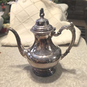 May include: A silver-colored teapot with a curved spout and handle. The teapot has a decorative lid with a small finial. The body of the teapot is rounded, and it sits on a small base. The teapot is reflective, showing the surrounding environment.