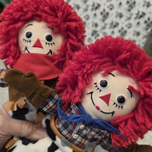 May include: Two Raggedy Ann and Andy dolls. The dolls have red yarn hair, embroidered faces, and are dressed in plaid shirts, blue bow ties, and cow-print pants. One doll wears a red bandana. The dolls are from the DAKIN Signature Collection.