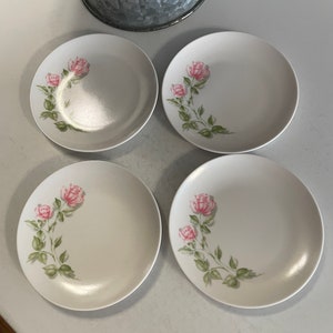 May include: Set of four white ceramic dessert plates with a pink rose floral design. The plates have a slightly raised rim and a smooth, glossy finish.