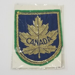 May include: A green and blue fabric patch with a gold embroidered maple leaf and the word "CANADA" in gold lettering.