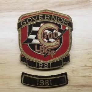 May include: A gold embroidered patch with the text "GOVERNOR 300 LOOP 1981" and a black and white checkered flag. The patch is on a wooden surface.