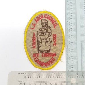 May include: Tan oval patch with yellow trim featuring a brown illustration of a person with a rifle, and the text "L.A. AREA COUNCIL BSA KIT CARSON CAMPOREE 1955". A ruler is next to the patch.