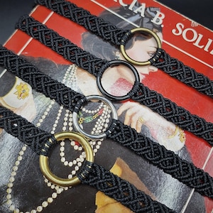 Puede incluir: Cuatro pulseras de macramé negro con intrincados patrones trenzados. Cada pulsera está centrada con un anillo metálico circular, incluyendo dos en tono latón, uno en tono plateado y uno en negro, todos asegurados con cordón negro.
