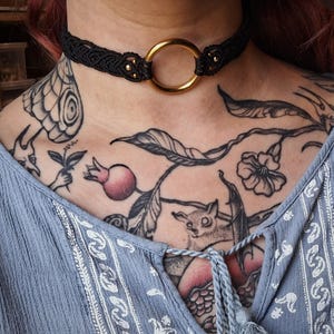 May include: Black macramé choker necklace with a gold-coloured ring and small gold beads. The necklace is worn on a person's neck. The person is wearing a blue top and has tattoos on their chest and shoulder.