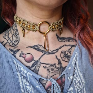 Peut inclure: Un collier ras du cou en macramé doré avec un grand anneau et un charm en forme d'épingle. Le choker présente de petites perles dorées et des accents pointus. Le collier est porté sur un haut bleu avec un motif floral.
