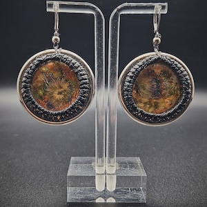 May include: A pair of round dangle earrings featuring a central resin inlay containing dried botanical elements in shades of orange, green, and brown. The circular resin pieces are framed by a black braided cord and a silver-toned metal border.
