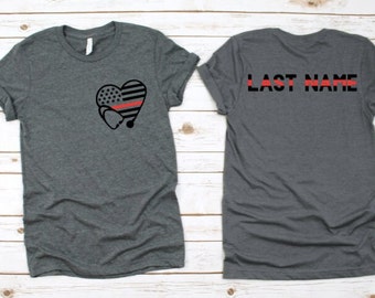 May include: A heather grey t-shirt with a black and red heart with a stethoscope and an American flag design. The back of the shirt has the text "LAST NAME" in black with a red line.