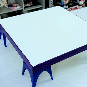 May include: A white canvas with a purple border is sitting on four blue easel legs. The canvas is ready for painting.