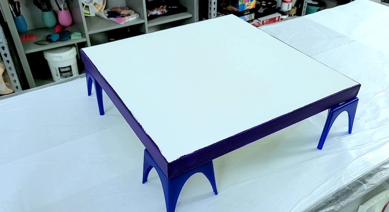 May include: A white canvas with a purple border is sitting on four blue easel legs. The canvas is ready for painting.