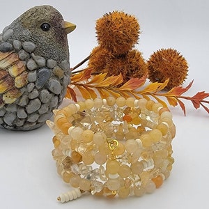 May include: A multi-strand bracelet made of light yellow and clear quartz chips and beads. The bracelet is wrapped around a decorative bird figurine.