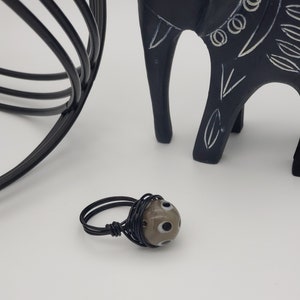 May include: A black wire wrapped ring with a brown and white glass bead. The bead has a pattern of white circles with black centers.