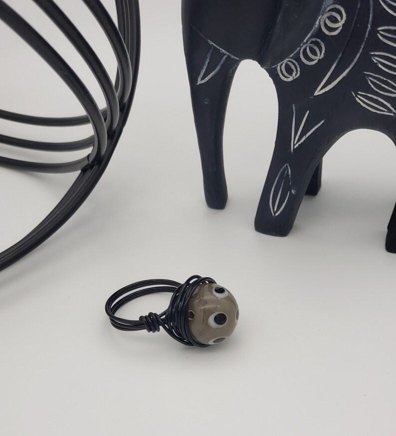 May include: A black wire wrapped ring with a brown and white glass bead. The bead has a pattern of white circles with black centers.