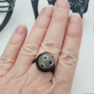 May include: A black wire wrapped ring with a gray and white patterned glass bead. The bead has a design of white circles on a gray background.