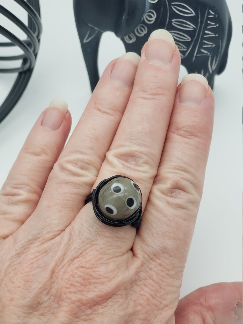 May include: A black wire wrapped ring with a gray and white patterned glass bead. The bead has a design of white circles on a gray background.