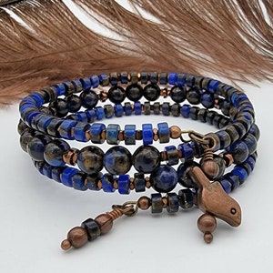 Könnte beinhalten: Ein mehrreihiges Armband mit blauen und braunen Perlen, einem Kupfervogel-Anhänger und einem Kupferperlen-Anhänger. Das Armband ist um eine Feder gewickelt.