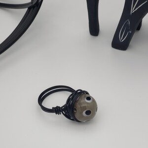 May include: A black wire wrapped ring with a gray and white bead featuring black eyes.