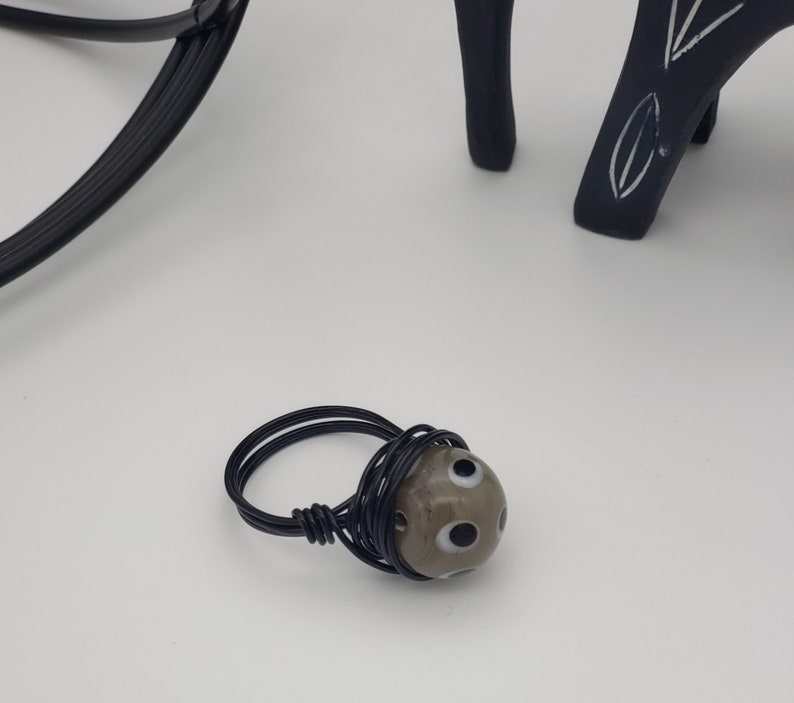 May include: A black wire wrapped ring with a gray and white bead featuring black eyes.