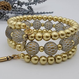 May include: A gold and white beaded bracelet with a geometric pattern. The bracelet is made of three strands of beads and has a gold charm.