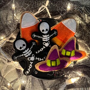 May include: A set of four Halloween-themed felt toys. Two are candy corn shaped, one is a witch's hat in purple and green, and one is a black skeleton.