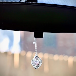 May include: A silver heart-shaped charm hangs from a silver chain attached to a rearview mirror in a car. The charm has a clear center with a pink tint.
