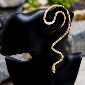 May include: A gold-toned ear cuff shaped like a long, winding snake that wraps around the outer edge of the ear. The entire surface of the snake is encrusted with small, clear, sparkling stones, and it features two tiny black stones for eyes.