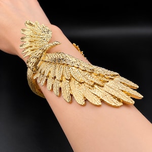Puede incluir: Un brazalete de metal en tono dorado con un diseño detallado de ala de águila que envuelve la muñeca. La superficie del ala muestra patrones de plumas intrincados y texturizados, con el ala principal extendiéndose a lo largo del antebrazo.