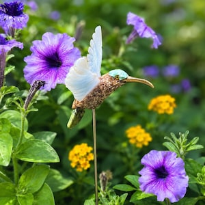 May include: A hummingbird garden stake featuring a textured brown body, translucent white wings, a turquoise head patch, and a long gold-toned beak. The bird is mounted on a thin metal rod, positioned in a garden with purple and yellow flowers.