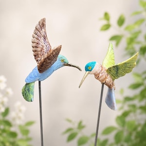 May include: Two hummingbird garden stakes featuring carved stone-like textures. The left bird has brown wings, a blue body, and a green tail. The right bird has a blue head, tan body, and light green wings. Both are mounted on thin metal rods.