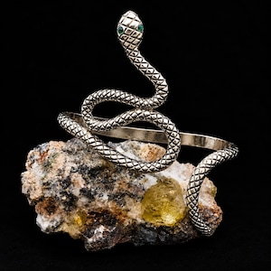 May include: A silver-toned metal cuff bracelet shaped like a coiled snake with a textured scale pattern and two small green stones set as eyes. The snake body forms the band of the bracelet, featuring a detailed head and a winding tail design.
