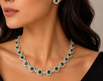 May include: A necklace and matching stud earrings set featuring round, vibrant green center stones surrounded by a halo of small, clear, sparkling white stones. The jewelry pieces are arranged in a floral-inspired pattern with silver-toned metal settings.