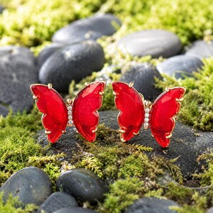 May include: A pair of butterfly-shaped stud earrings featuring vibrant red faceted wings and a central vertical row of small clear stones set in a gold-toned metal frame, showcasing a detailed wing silhouette and a polished, reflective finish.