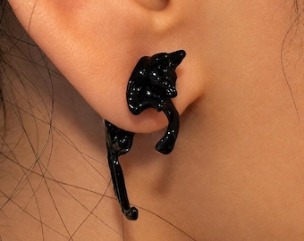 May include: A glossy black earring shaped like a cat, designed to wrap around the earlobe. The cat figure features a smooth, reflective finish with the head positioned at the top and the tail extending downward along the side of the earlobe.