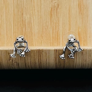 May include: A pair of silver frog earrings. The frogs are sitting on a wooden surface.