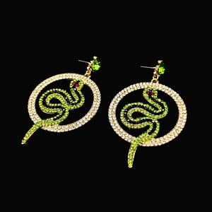 May include: A pair of gold-toned snake earrings. Each earring features a green snake design within a crystal-studded oval frame. The snake has red eyes and a green gem at the top. The background is black.