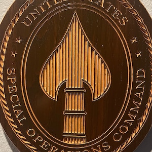 May include: A circular wooden plaque, featuring the words "UNITED STATES SPECIAL OPERATIONS COMMAND" carved around the edge. The centre has a detailed spearhead design. The plaque is a warm brown colour.