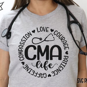 May include: A gray t-shirt with a black circle design featuring the text "CMA life" surrounded by the words "Compassion", "Love", "Courage", "Patience", and "Caffeine". A stethoscope is visible on the shirt and around the model's neck.