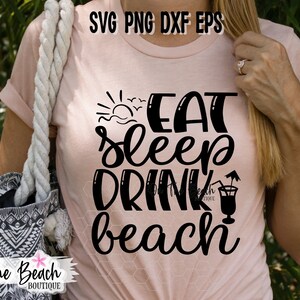 May include: A pink t-shirt with the text "Eat Sleep Drink Beach" in black lettering. The text is in a cursive font. A sun, waves, and seagulls are above the text. A cocktail glass with an umbrella is next to the text.